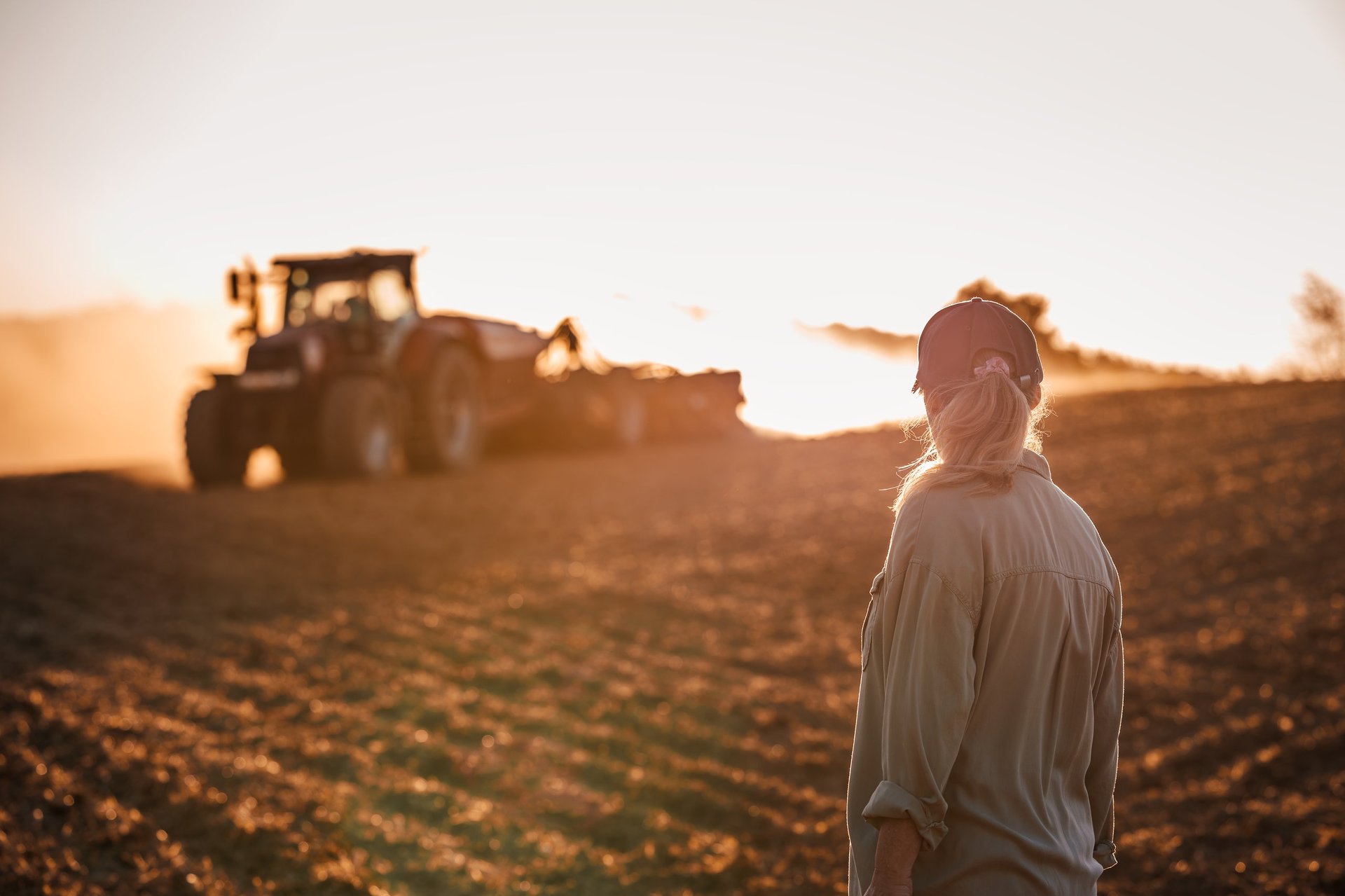 Farmer supervising tractor during agriculture sowing in field. Woman agronomist looking at agricultural activity during sunset