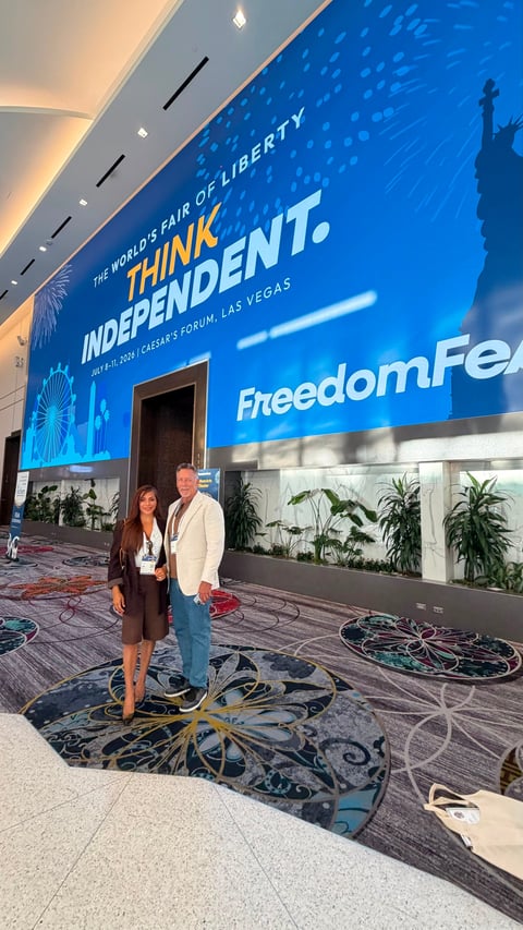 Two people standing in front of a large blue billboard for FreedomFest at the Caesars Forum in Las Vegas