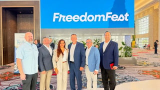 Group of six business professionals posing together under FreedomFest banner in hotel lobby