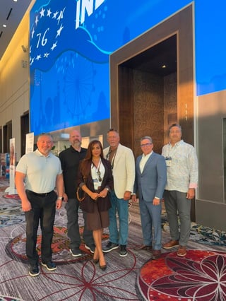 Six business professionals standing together in an indoor venue with blue patriotic signage and decorative red carpet