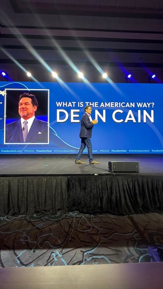 Speaker on stage in dark suit presenting at political event with Dean Cain image and What is the American Way? text on blue screen