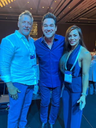Three people smiling at an indoor event, wearing blue clothing and lanyards, standing in front of a modern ceiling with recessed lighting