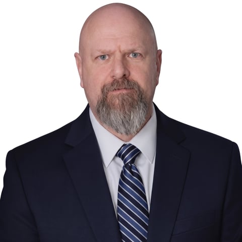 Professional headshot of a bald man with a gray beard wearing a dark blazer, light blue shirt, and striped tie against a white background.