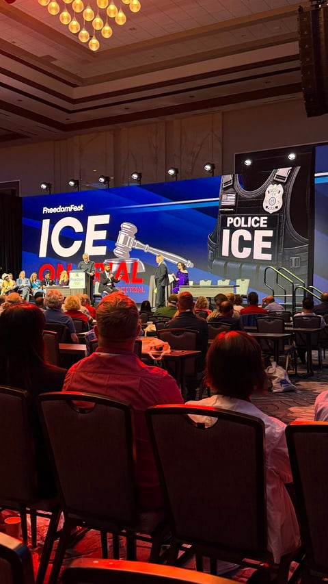 Audience seated in a conference hall watching a stage presentation with ICE and Police ICE branded screens and a microphone setup.