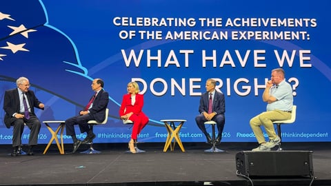 Panel discussion on stage with four speakers seated in chairs discussing American achievements, with blue backdrop and American flag design.